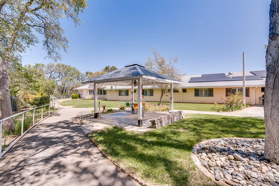 Outdoor area of Meadowood Nursing Center featuring a paved walkway, a small covered pavilion with seating, green grass, trees, and a single-story building with solar panels on the roof under a clear blue sky.