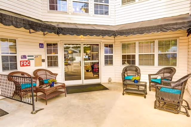 Entrance patio of a senior living facility with wicker chairs and teal cushions arranged around a glass double door under a black awning.
