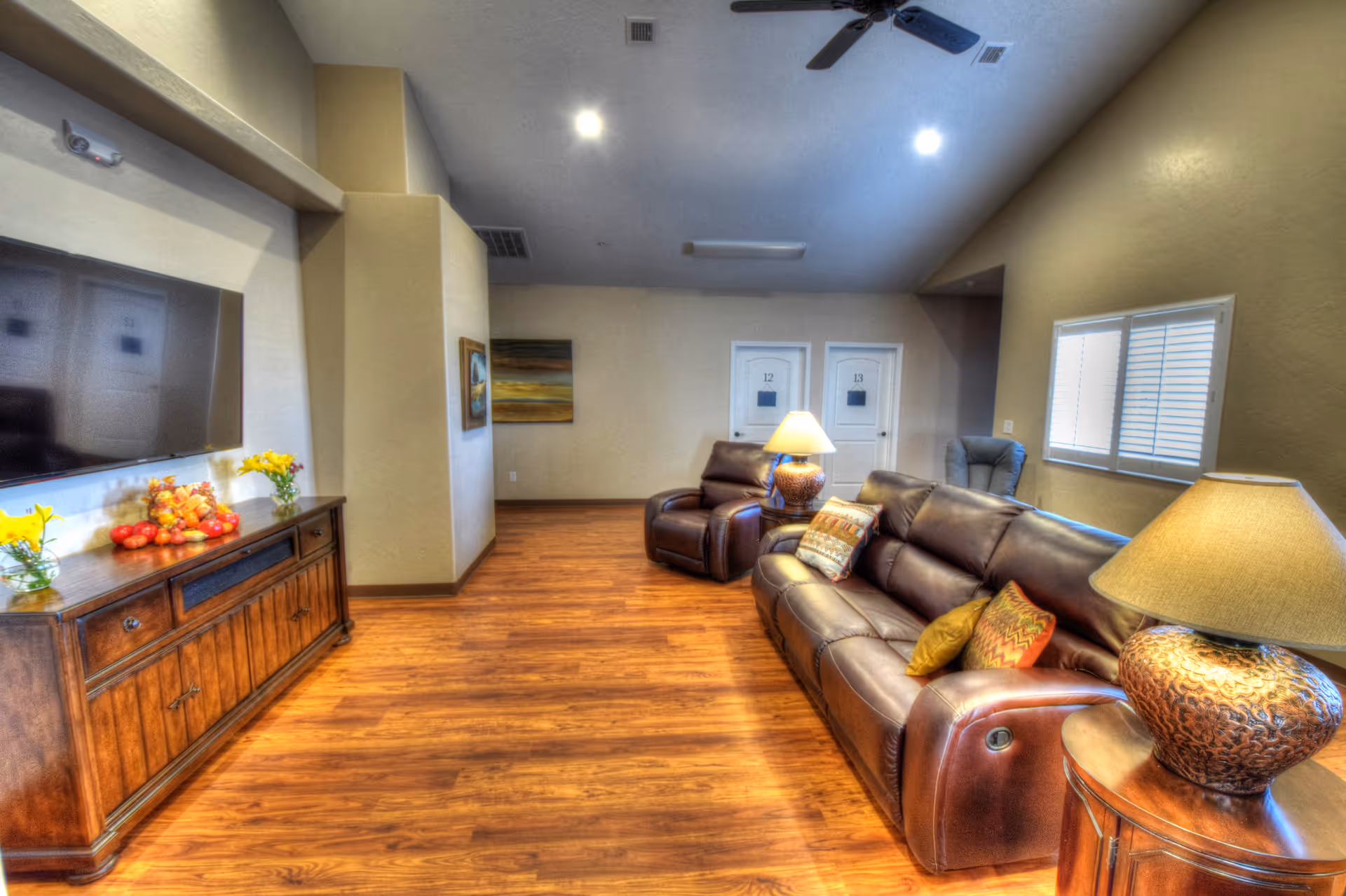 A cozy living room with wooden flooring, a large brown leather sofa with colorful cushions, a matching leather recliner, two table lamps with textured bases, a wooden cabinet with decorative items including flowers and fruit, a large flat-screen TV mounted on the wall, and two white doors labeled 12 and 13 in the background.