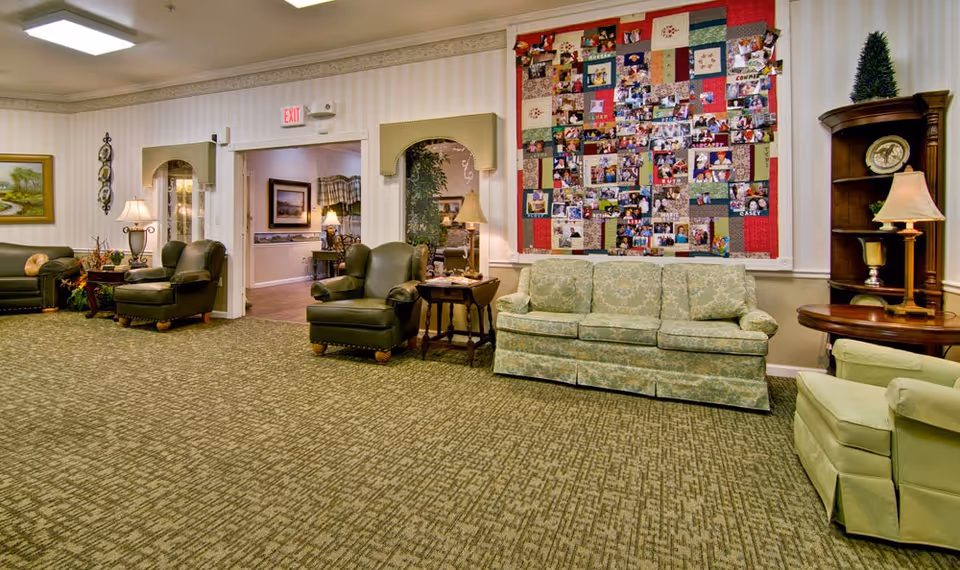 A cozy senior living common area with green upholstered armchairs and a matching sofa arranged around small wooden side tables. The room features a patterned carpet and light-colored walls with framed artwork. A large colorful quilt with photos is displayed on the wall. A wooden corner shelf with a lamp and decorative items is also visible.