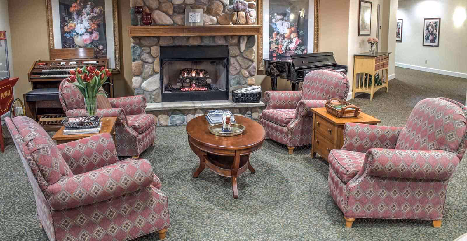 A cozy living room area with four patterned armchairs arranged around a wooden coffee table. The room features a stone fireplace with a fire burning, a piano, an organ, floral paintings on the walls, and a small wooden side table with a basket and flowers.