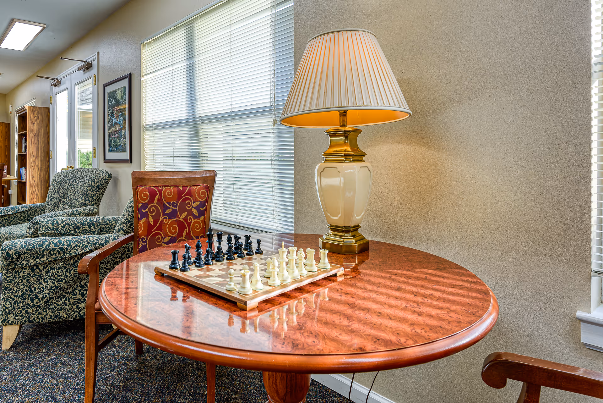 Round wooden table with a chessboard and lamp in a cozy seating area with armchairs and windows.