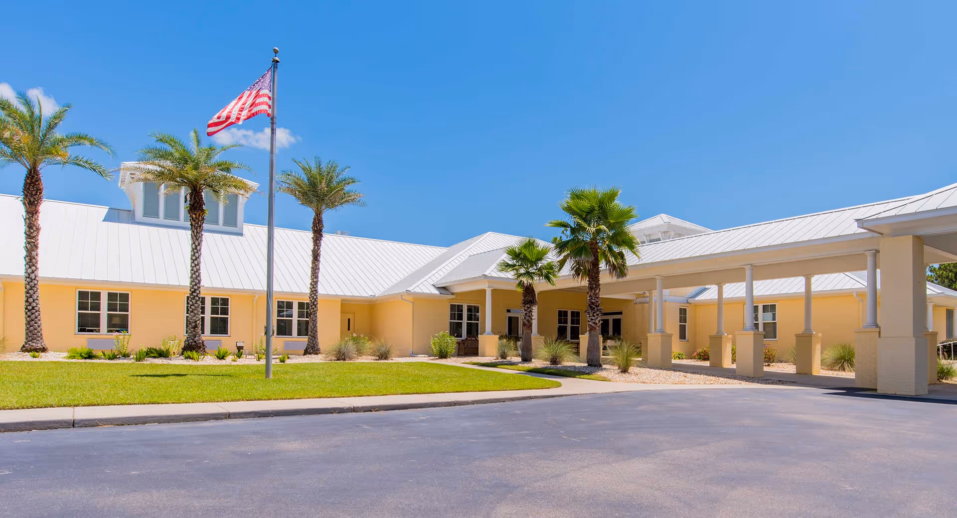 Single-story yellow memory care building with a covered entrance, palm trees, a flagpole, and a clear blue sky.