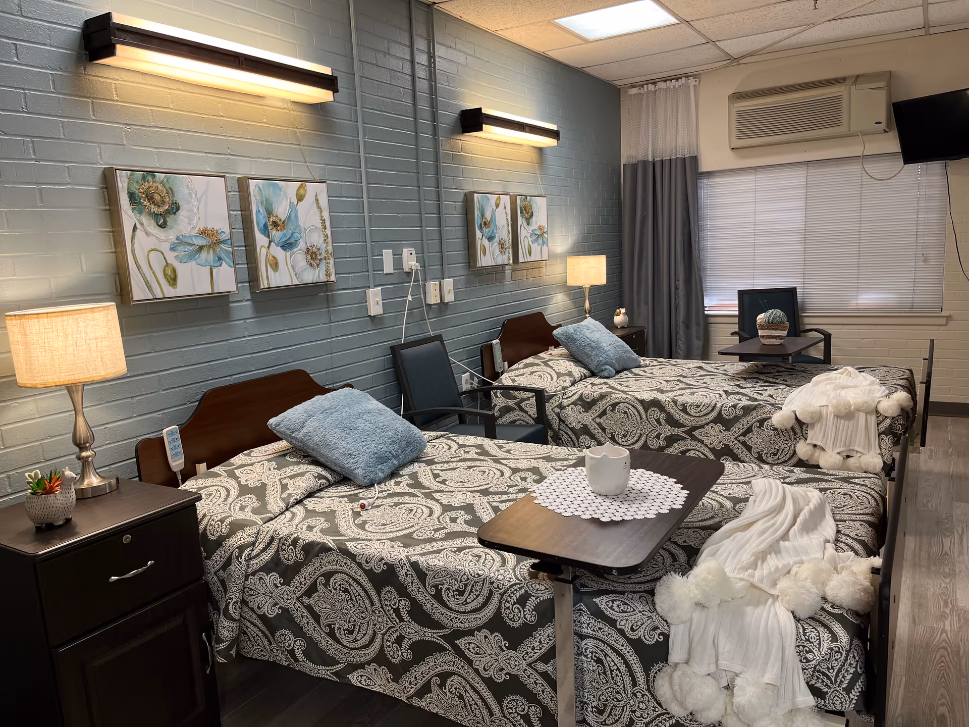 Two neatly made twin beds in a nursing facility room with nightstands, lamps, and wall art.