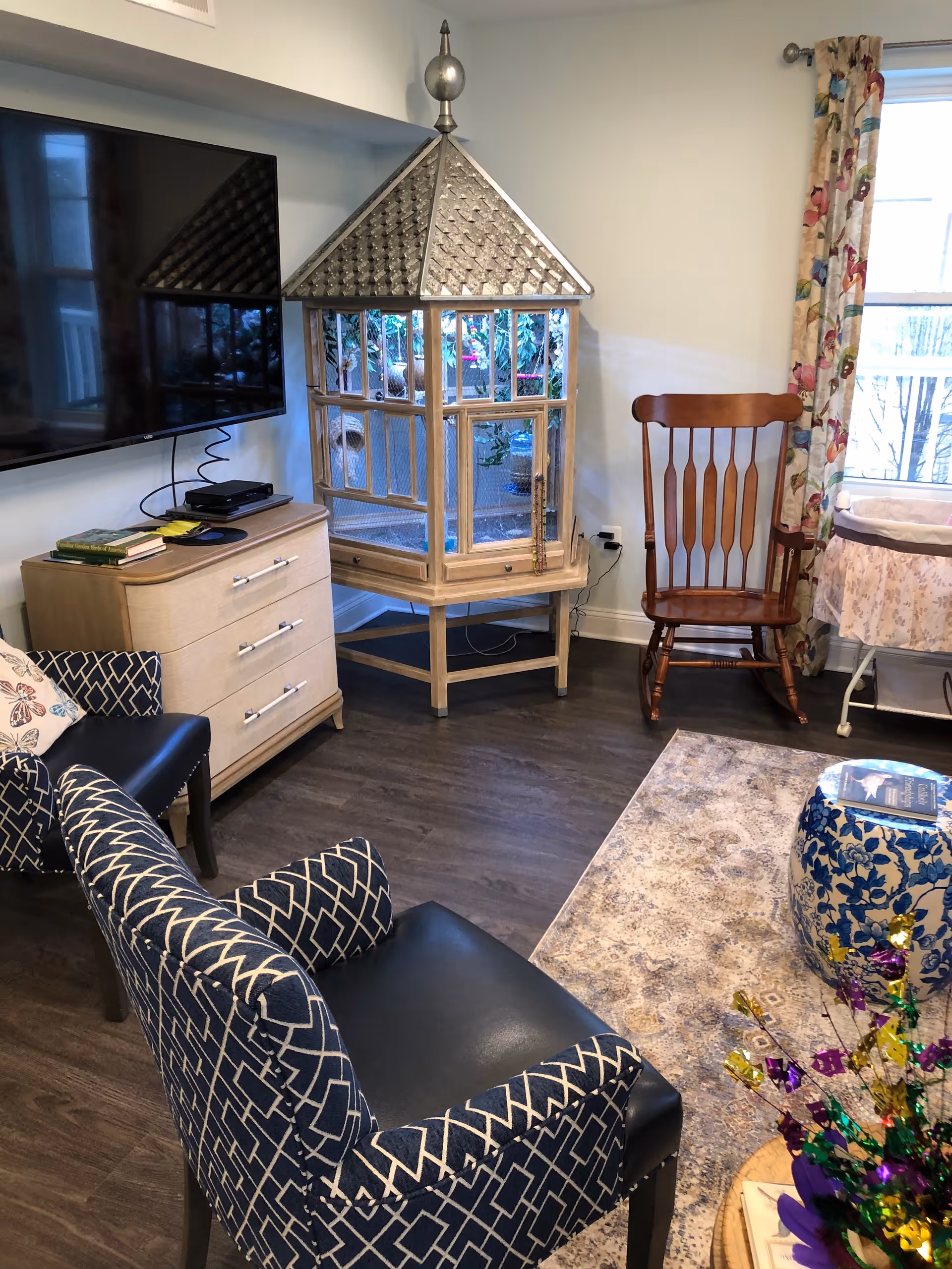 A cozy living room area with two patterned armchairs, a wooden rocking chair, a large flat-screen TV mounted on the wall, a light wood dresser with books and electronic devices on top, a decorative birdcage, a floral curtain covering a window, and a patterned rug on the floor.