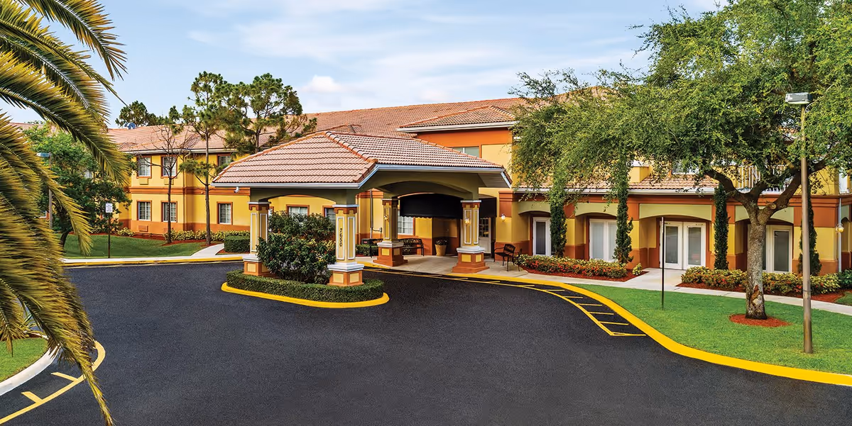 Exterior view of Wickshire Tamarac senior living facility showing a covered entrance with a tiled roof, surrounded by landscaped greenery, trees, and a paved driveway with yellow curbs.