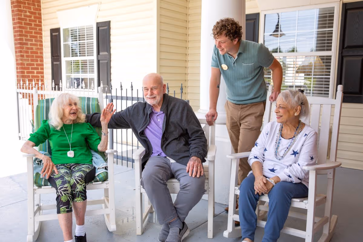 Three elderly individuals sitting on rocking chairs on a porch, smiling and interacting with a young male caregiver who is standing and leaning slightly towards them. The porch has light-colored siding, a white column, and a window with black shutters in the background.