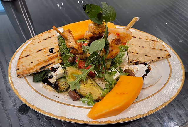 A plate with grilled shrimp skewers, slices of flatbread, avocado, olives, arugula, feta cheese, and two slices of cantaloupe melon arranged on a white plate with a gold rim, placed on a dark glass table.