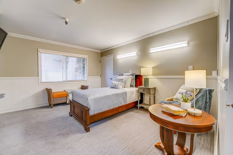 A neatly arranged bedroom in a memory care facility featuring a single wooden bed with white and patterned pillows and a light gray bedspread. The room has beige walls with white wainscoting, a window with blinds, a small bench under the window, a bedside table with a lamp, and a round wooden table with a lamp, books, and a small plant. There is also a cushioned chair with a throw blanket and pillow next to the round table.