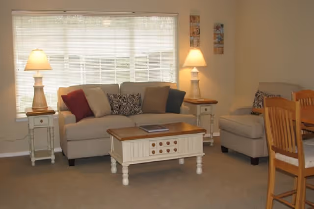 A cozy living room with a beige sofa adorned with multiple cushions, two matching side tables each with a lamp, a white wooden coffee table with a book on it, a beige armchair, and a wooden dining table with chairs. A large window with blinds is behind the sofa, allowing natural light into the room.