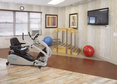 Indoor fitness room with elliptical trainer, small wooden stairs, exercise balls, and a wall TV.