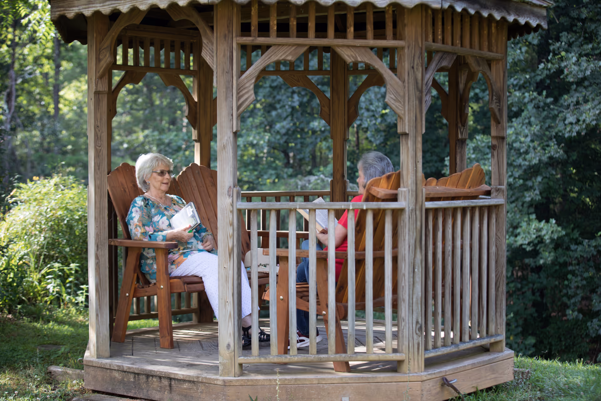 Two elderly women sitting and reading in wooden chairs inside a wooden gazebo surrounded by greenery and trees.