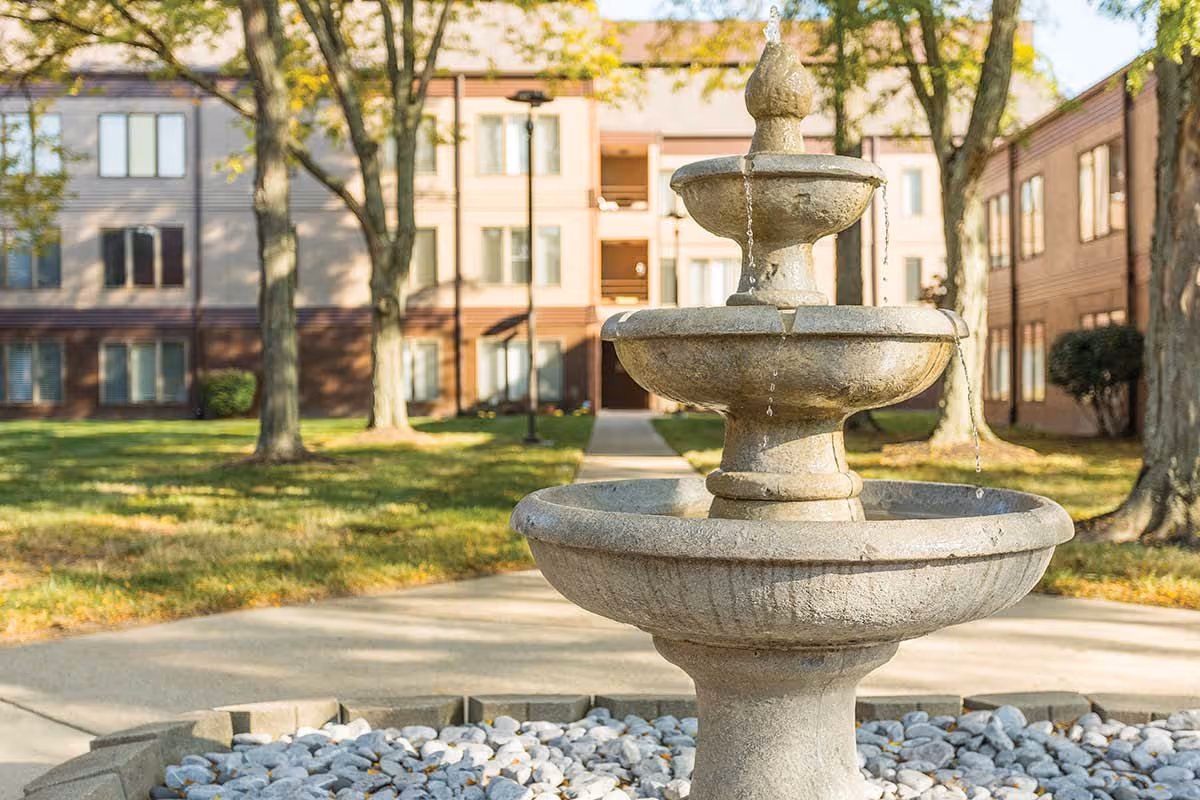 Tiered stone fountain in a grassy courtyard in front of a multi-story senior living building.