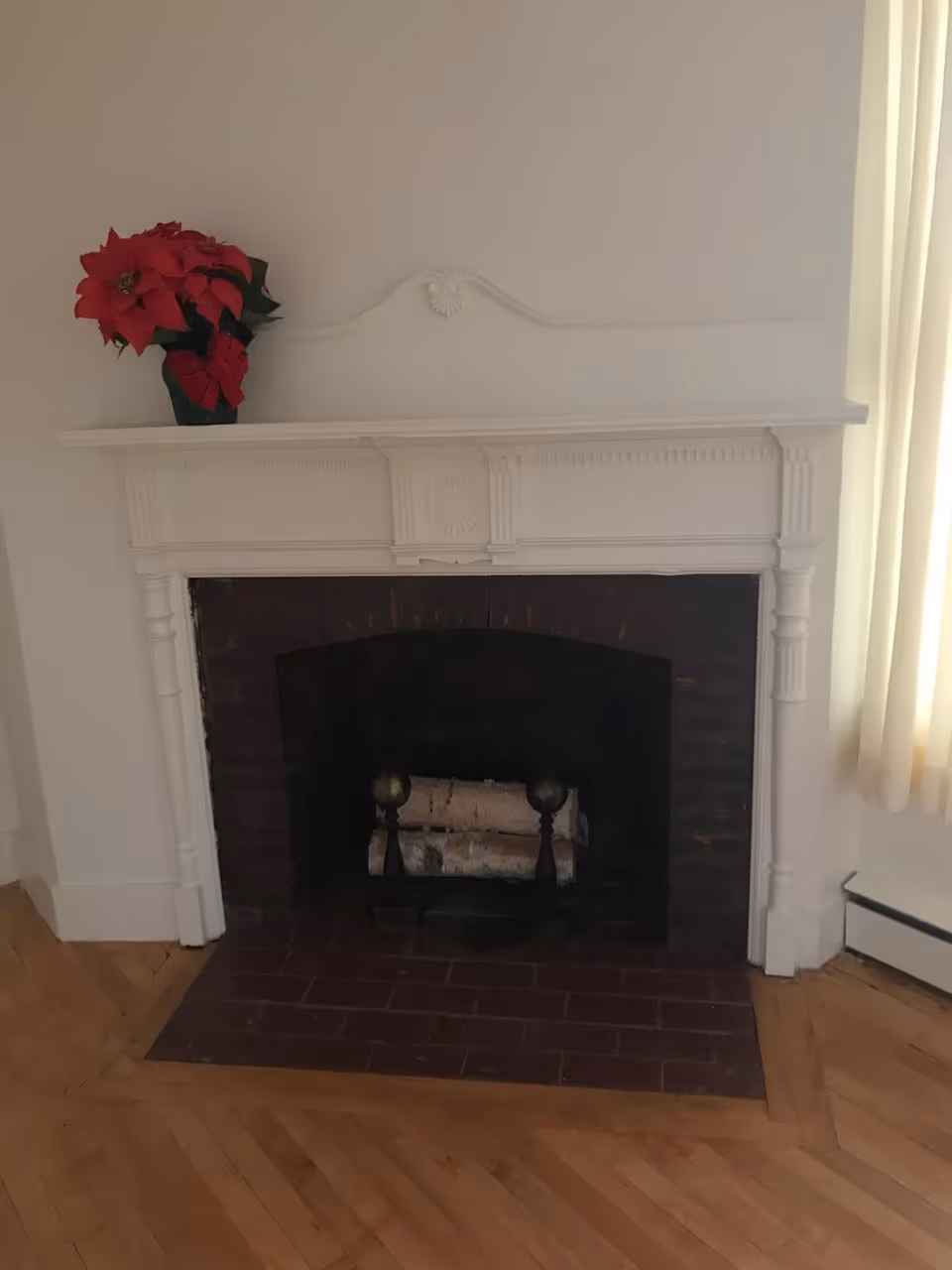A white decorative fireplace with a dark brick interior and two logs placed inside. On the mantel, there is a red poinsettia plant in a black pot. The floor is wooden with a herringbone pattern, and there is a cream-colored curtain to the right side of the fireplace.