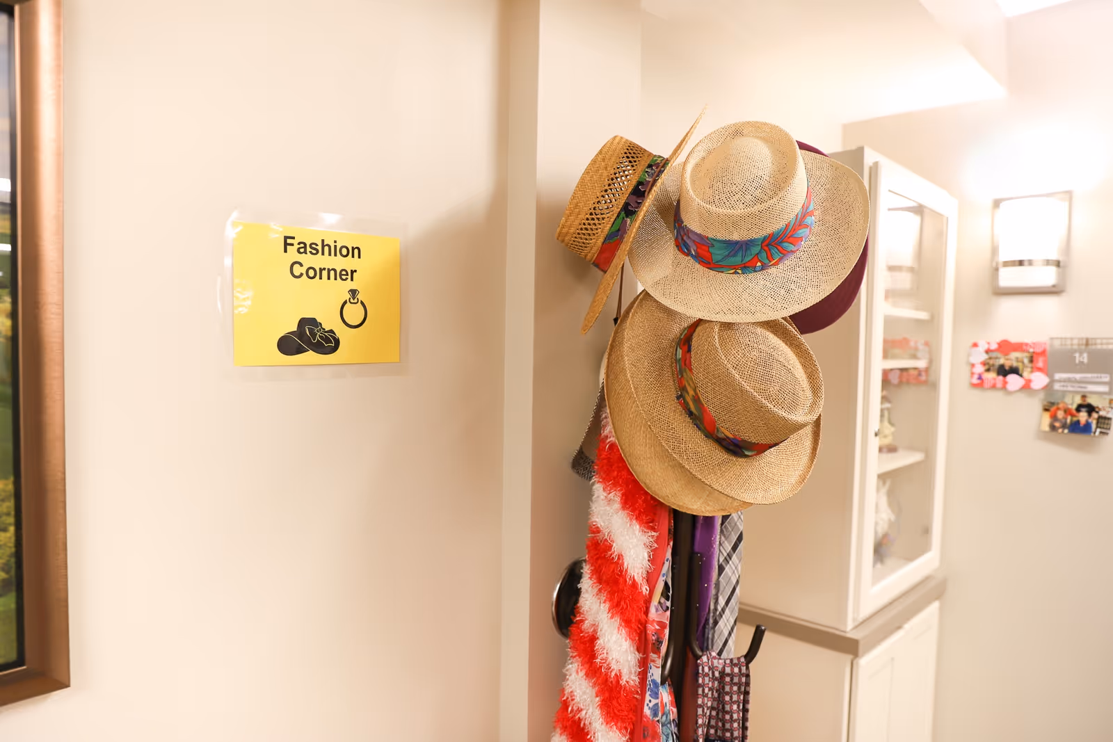 A corner labeled 'Fashion Corner' with a yellow sign on the wall. Several straw hats with colorful bands hang on a coat rack, along with scarves and ties. The area appears to be inside a senior living facility hallway with a light fixture and a bulletin board in the background.