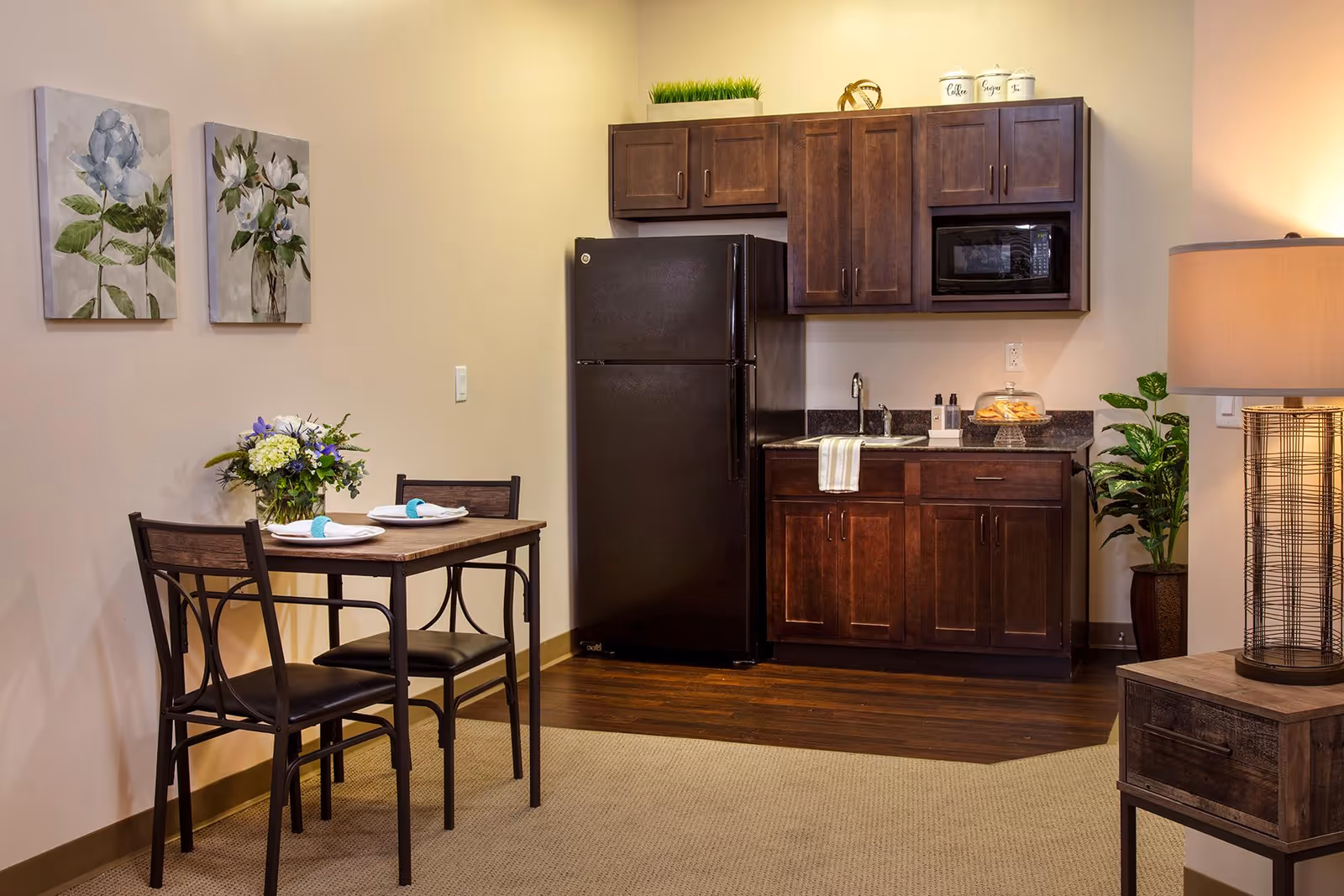 A small kitchen area with dark wooden cabinets, a black refrigerator, a microwave, and a sink. Next to the kitchen is a small dining table set for two with plates and napkins, and a vase of flowers. The room has light-colored walls with two floral paintings, a potted plant, and a lamp on a wooden side table.