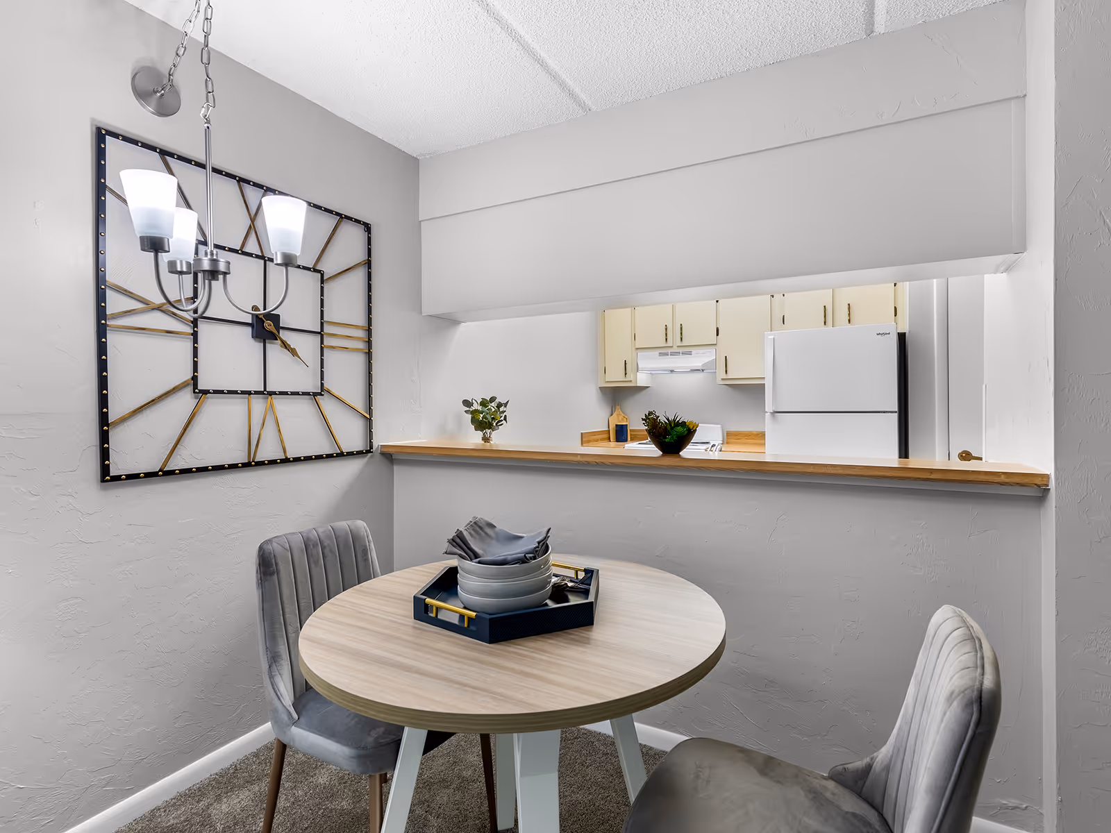 Small dining nook with a round table, two upholstered chairs, decorative wall clock art, and a pass-through counter to the kitchen.