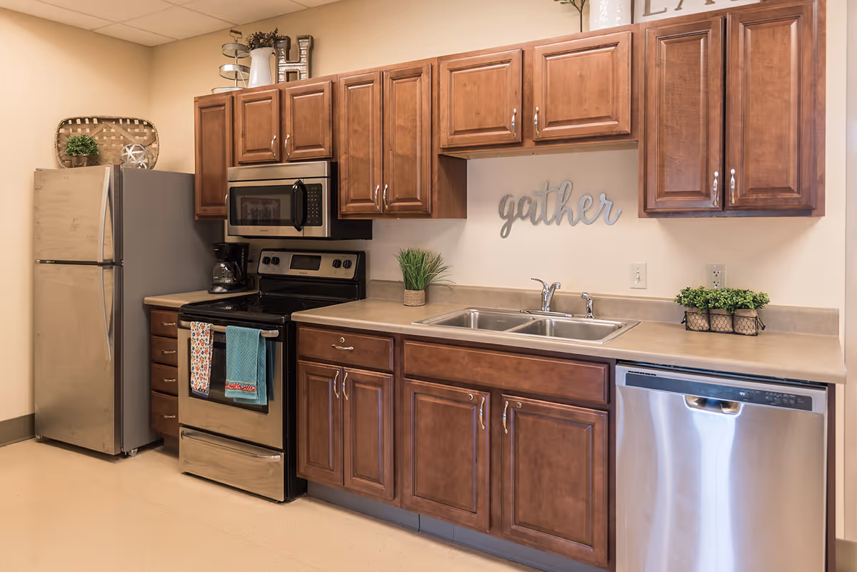 A modern kitchen with stainless steel appliances, wooden cabinets, a double sink, and a decorative 'gather' sign above the countertop.