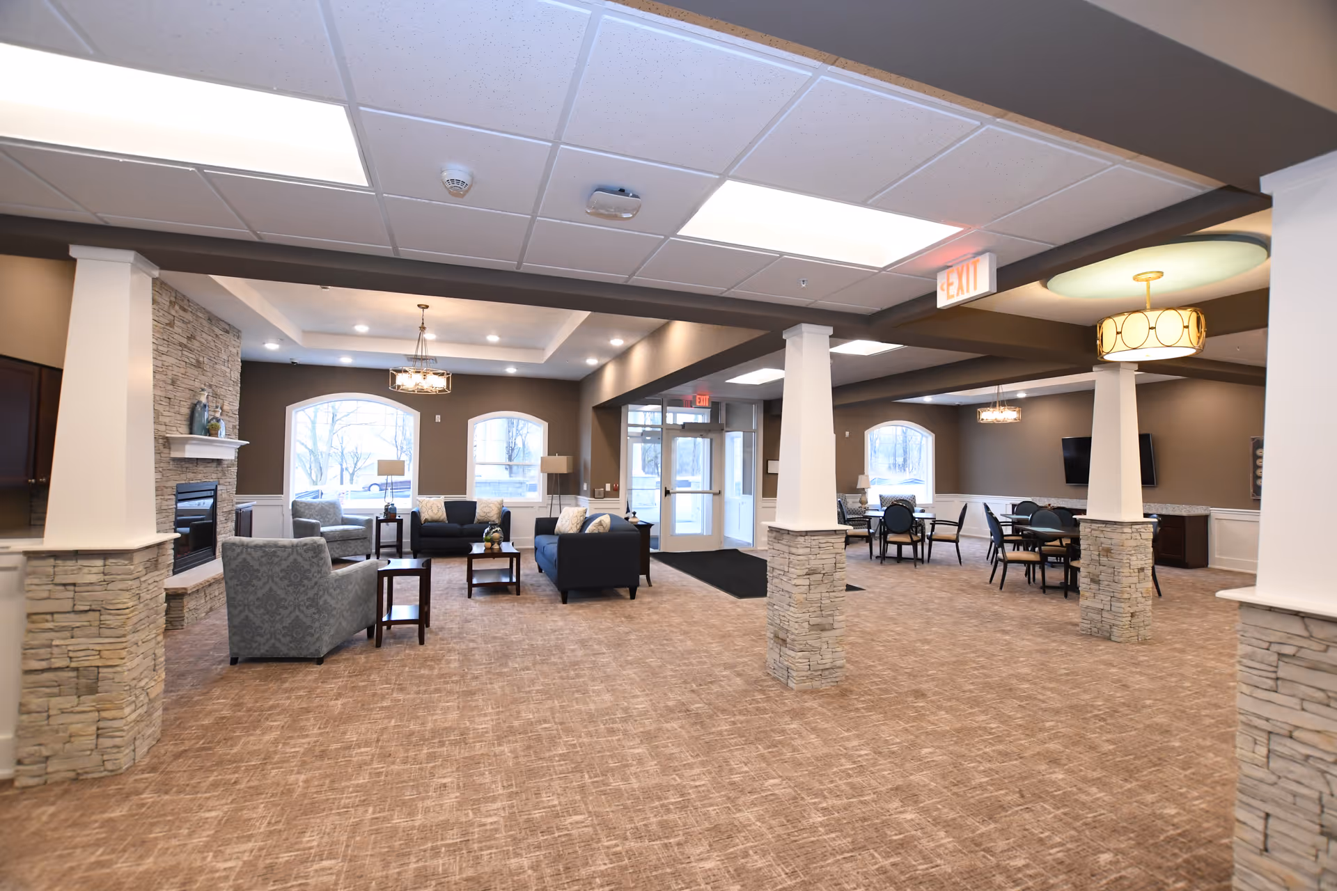 Spacious furnished common lounge with sofas, armchairs, tables, stone-clad columns, a fireplace and entry doors.