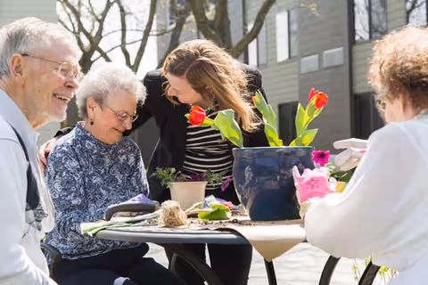 A group of elderly people and a caregiver are gathered around a table outdoors, planting flowers in pots. They are smiling and enjoying the activity on a sunny day with trees and a building in the background.