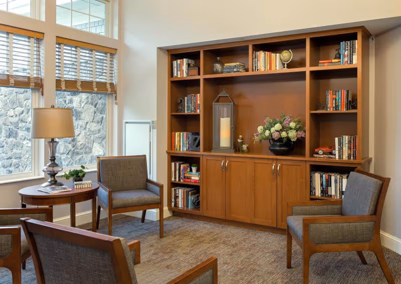 A cozy sitting area in a senior living facility with four wooden armchairs upholstered in gray fabric arranged around a wooden side table with a lamp and a small plant. Behind the chairs is a large wooden bookshelf filled with books, decorative items, a large candle lantern, and a flower arrangement. Large windows with wooden blinds let in natural light and show a stone wall outside.