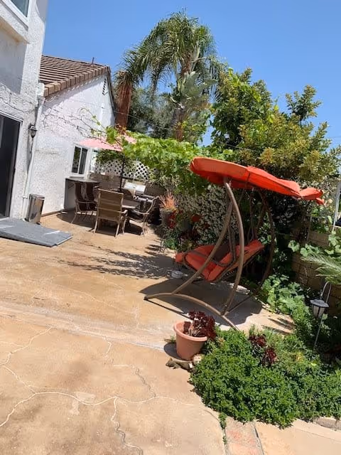 Outdoor patio area with a red cushioned swing chair under a red canopy, surrounded by green plants and trees. There is a dining table with chairs and a pink umbrella near a white building with a tiled roof. The sky is clear and blue.