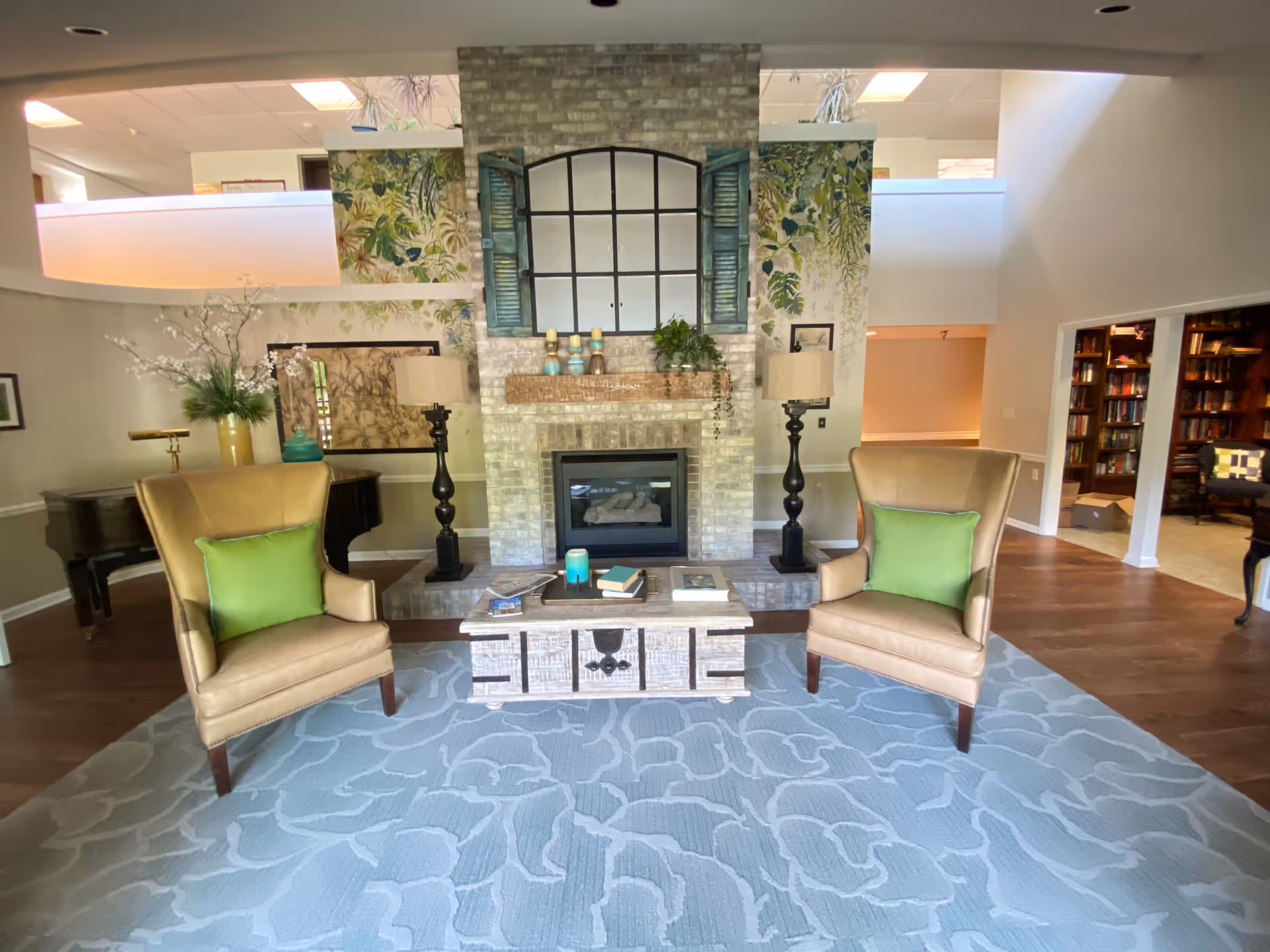 Cozy communal living room with a central brick fireplace, two tan armchairs with green pillows, and a coffee table on a patterned rug.