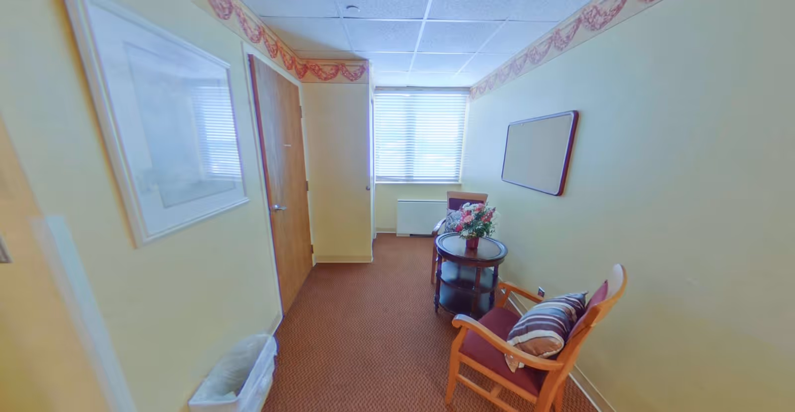 Small sitting room with a wooden armchair, round side table holding flowers, a window at the far end and a doorway on the left.