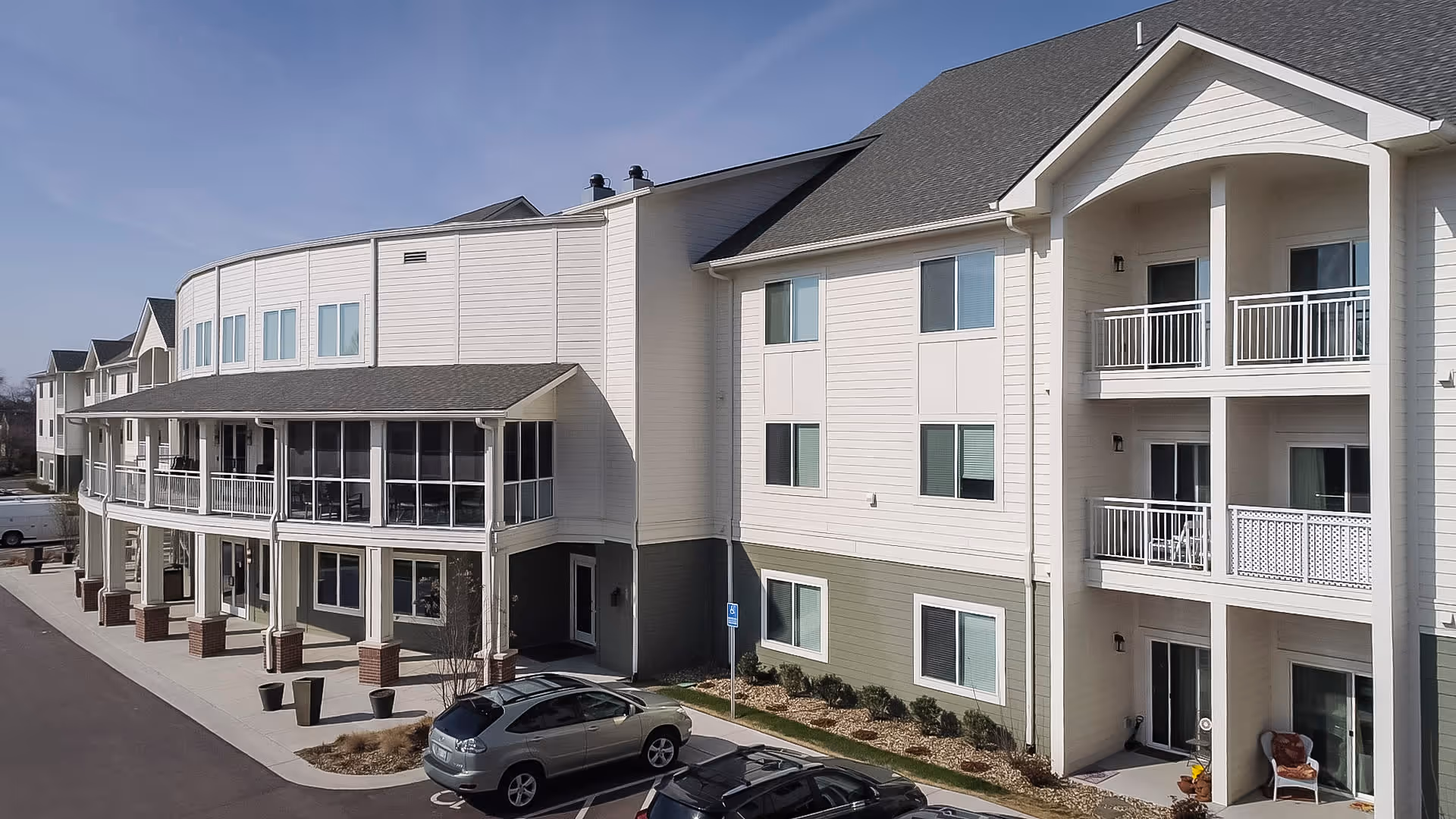 Exterior view of a multi-story assisted living facility building with balconies, large windows, and a covered entrance. There are a few parked cars in front of the building and a clear blue sky above.