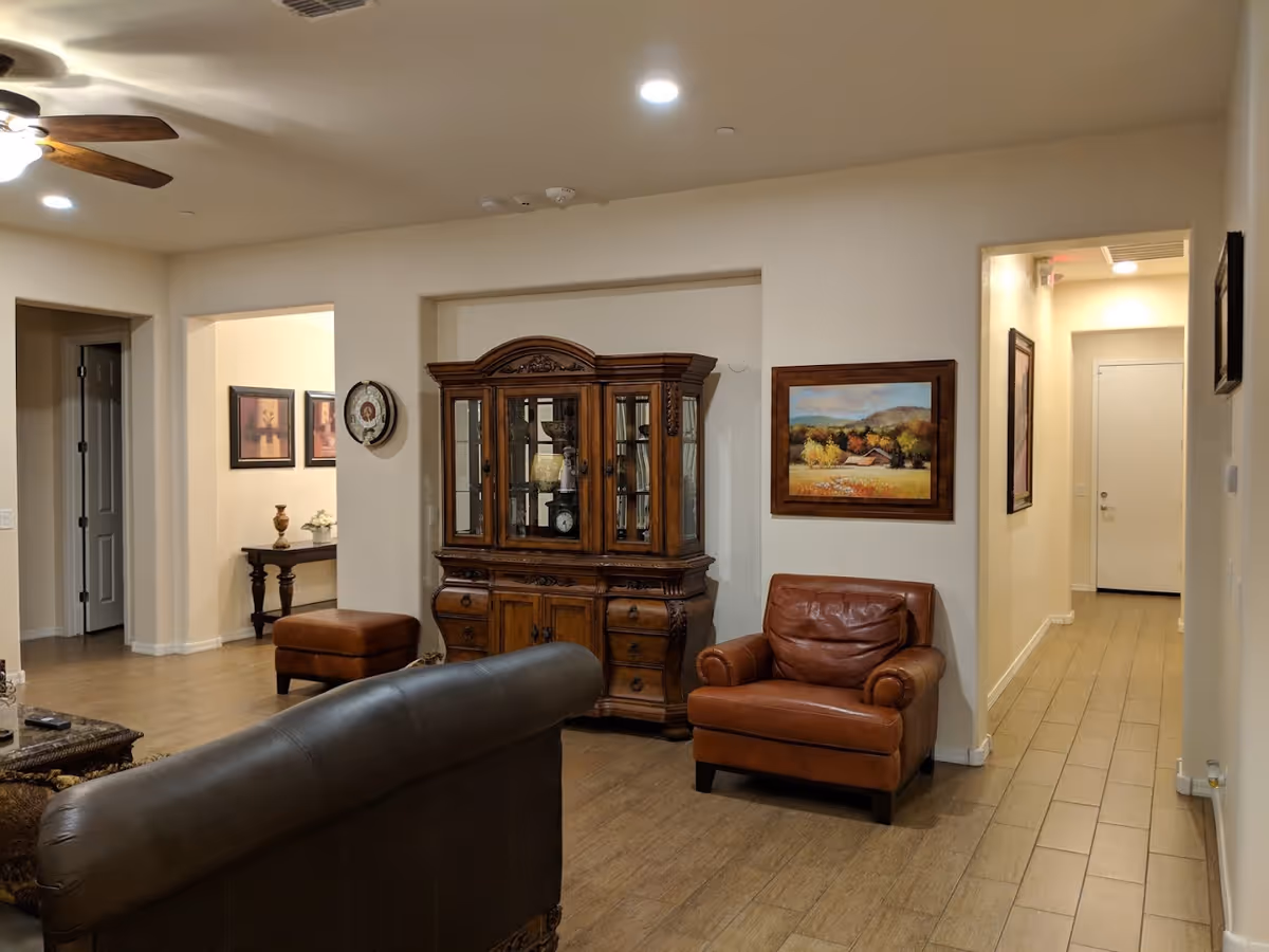 A cozy living room area with a brown leather armchair and a dark leather sofa. There is a wooden display cabinet with glass doors against the wall, and a framed landscape painting hanging beside it. The room has wooden flooring and recessed ceiling lights, with a ceiling fan visible. There are doorways leading to other rooms and a hallway with a white door at the end.