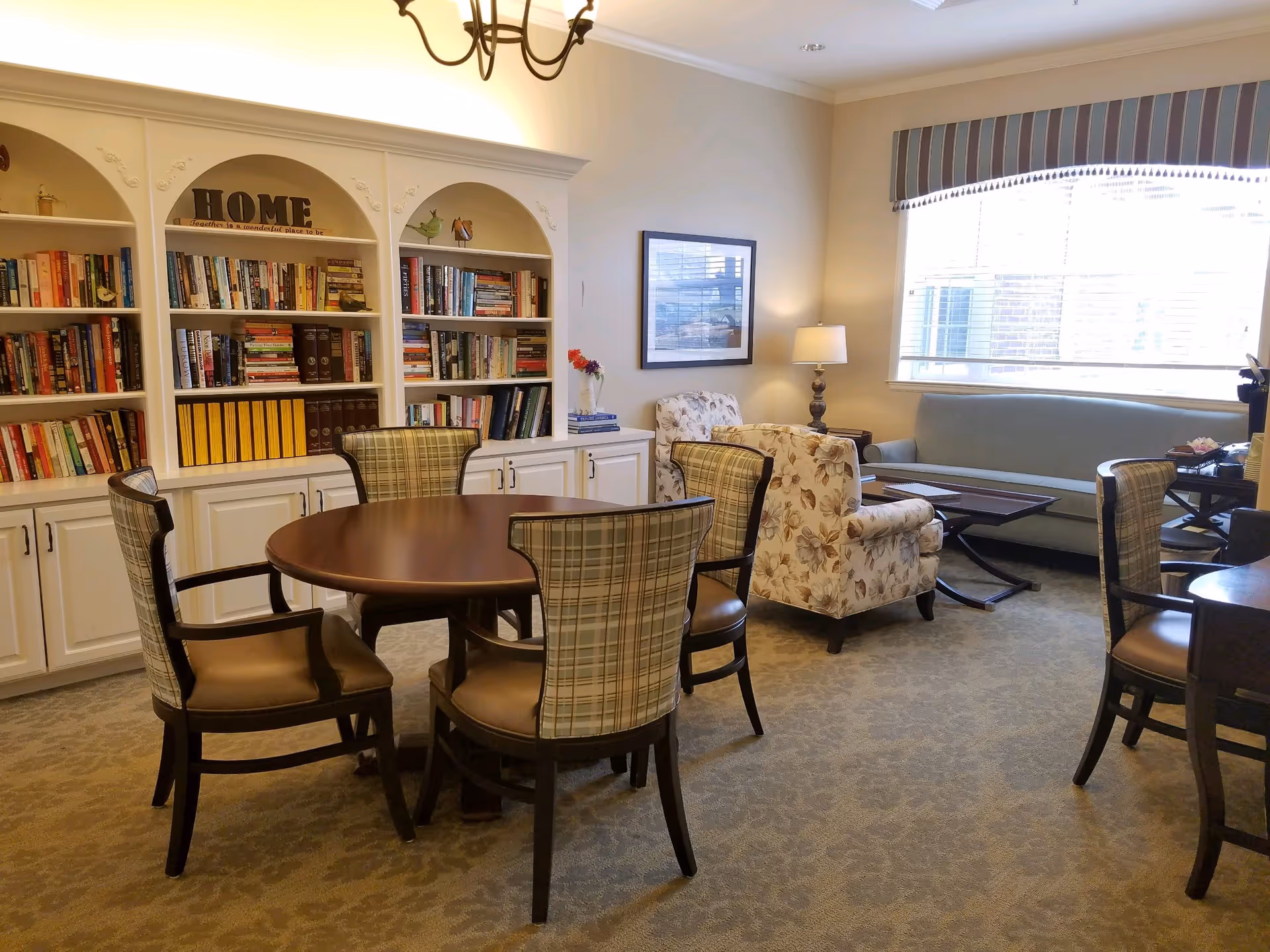 A cozy living room area with a round wooden table surrounded by four plaid upholstered chairs. Behind the table is a white built-in bookshelf filled with books and decorative items, including a sign that says 'HOME'. To the right, there are two floral armchairs, a floor lamp, a framed picture on the wall, and a large window with striped valance letting in natural light. A blue sofa and a coffee table are also visible near the window.
