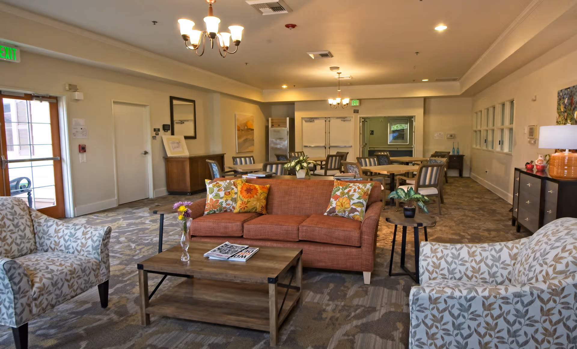 A spacious and well-lit common area in a senior living facility featuring a brown sofa with floral cushions, two patterned armchairs, a wooden coffee table with a vase of flowers and magazines, several dining tables with chairs in the background, and decorative plants and lamps. The room has carpeted floors, chandeliers, and exit doors visible.