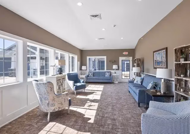 Bright senior living facility lounge with blue sofas, armchairs, lamps, and large windows.