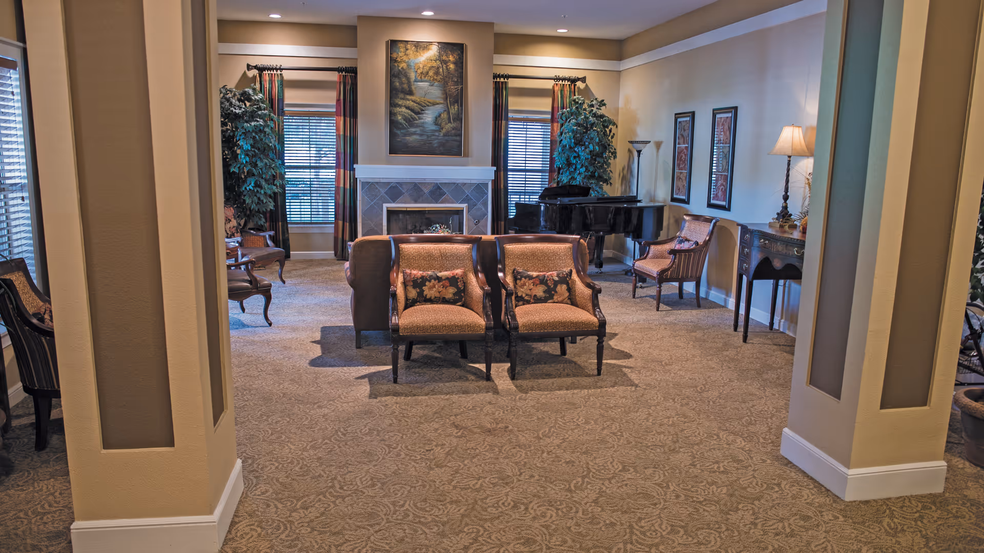 A cozy living room area with patterned carpet, two upholstered armchairs with floral cushions facing away, a fireplace with a landscape painting above it, two large windows with colorful curtains, a grand piano in the corner, and additional chairs and a small table with a lamp along the walls.