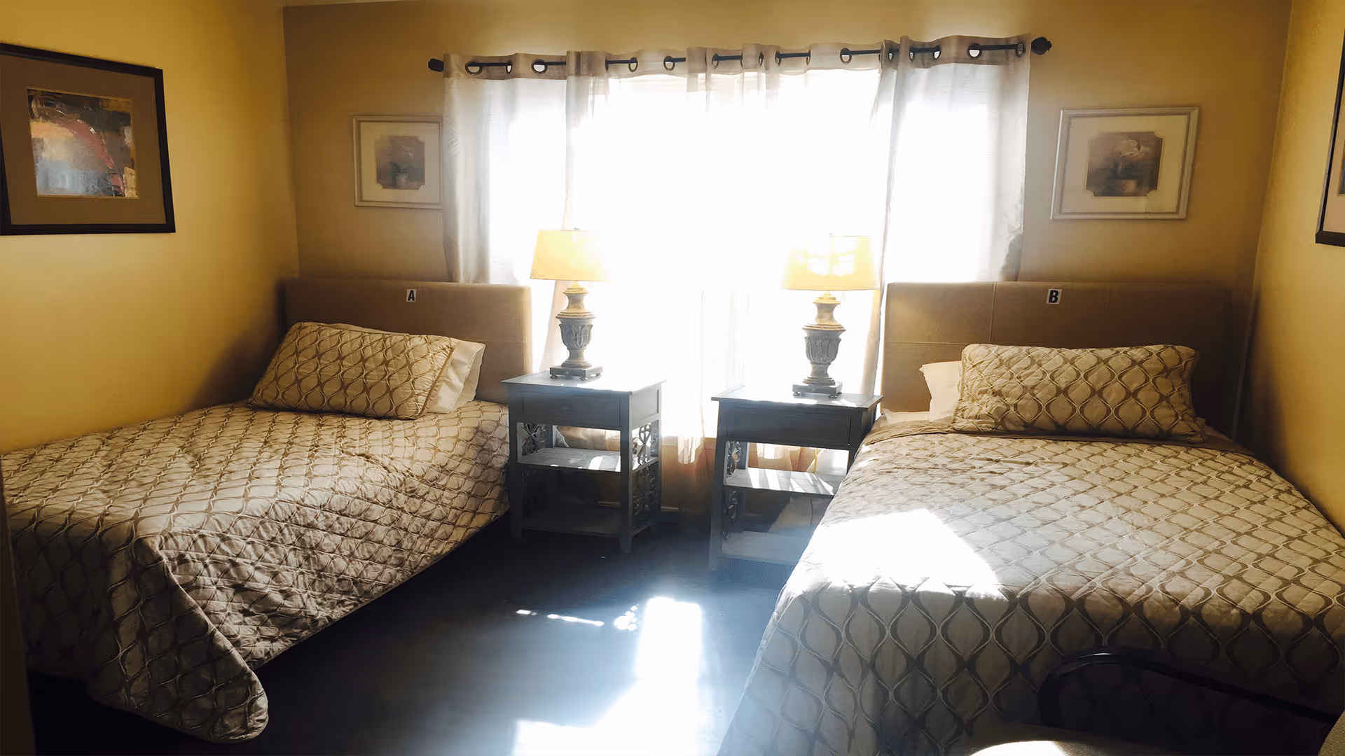 A cozy bedroom with two single beds separated by two small nightstands, each with a lamp. The beds have matching beige patterned bedspreads and pillows. There is a window with sheer curtains behind the nightstands, allowing sunlight to brighten the room. The walls are painted a warm yellow and decorated with framed artwork.