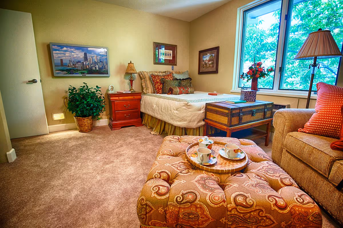 A cozy bedroom in a retirement community featuring a bed with multiple decorative pillows, a wooden nightstand with a lamp, a large window with a view of green trees, a patterned ottoman with a tray holding three teacups and saucers, a sofa with red cushions, a floor lamp, a framed cityscape painting on the wall, and a potted plant in a wicker basket.