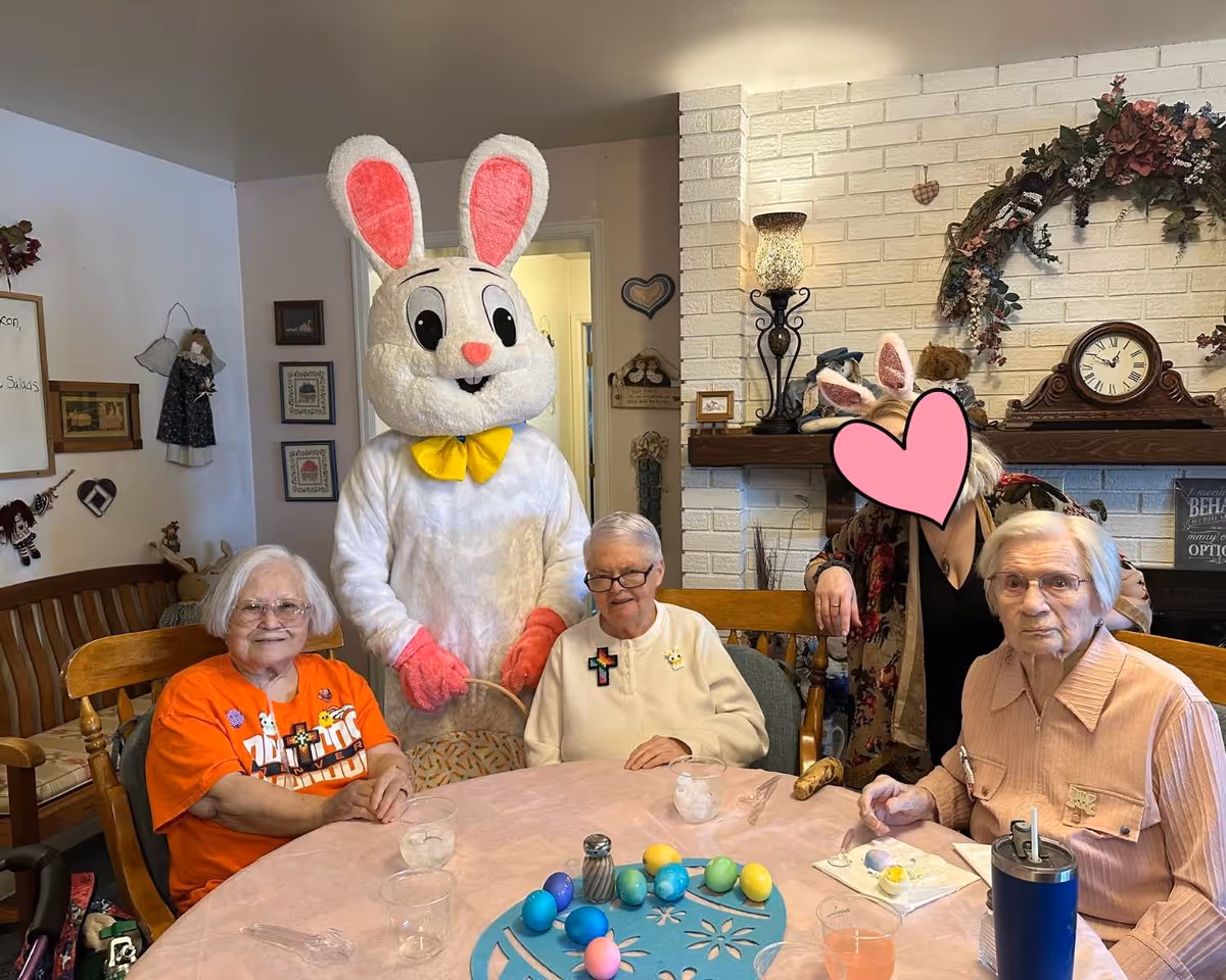 Three elderly residents sit around a table with colorful painted Easter eggs while a person in an Easter bunny costume and another adult pose behind them in a decorated common room.
