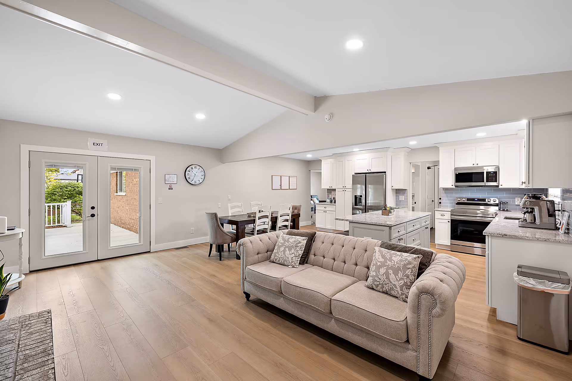 A bright and modern open-concept living area featuring a beige tufted sofa with patterned cushions, a wooden dining table with chairs, and a kitchen with white cabinetry, stainless steel appliances, and a central island. Double glass doors lead outside, and a wall clock is visible above the dining area.
