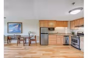 A bright kitchen and dining area featuring wooden cabinets, stainless steel appliances including a refrigerator, dishwasher, and stove. There is a small wooden dining table with two chairs, a painting on the wall, and a lamp on a side table in the background. The floor is light-colored hardwood.