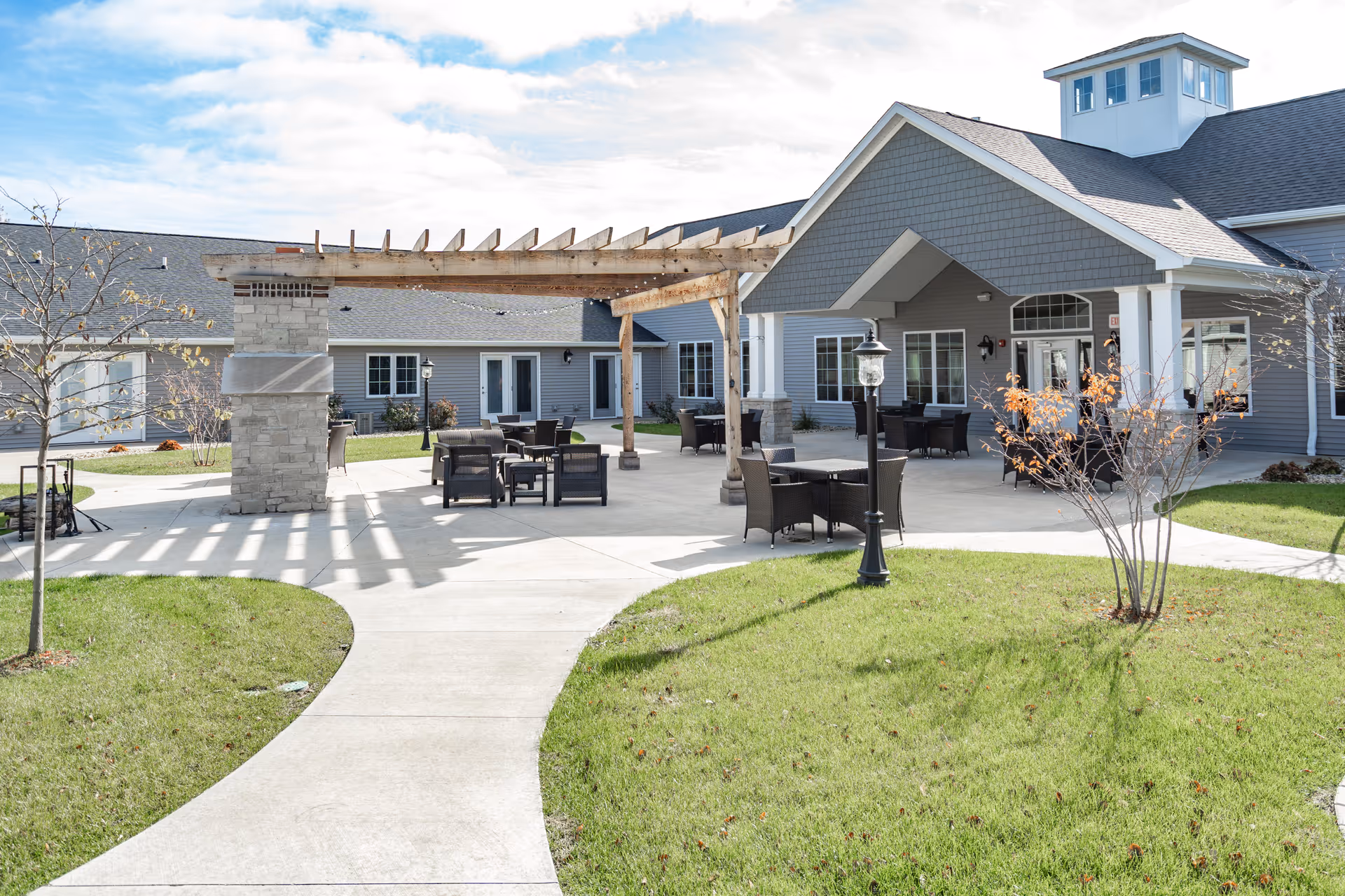 Outdoor patio area at Villas of Holly Brook Assisted Living & Memory Care in Pekin, IL, featuring a paved walkway, green grass, leafless trees, a wooden pergola with stone pillars, several black wicker chairs and tables, and a gray building with a covered entrance under a partly cloudy sky.