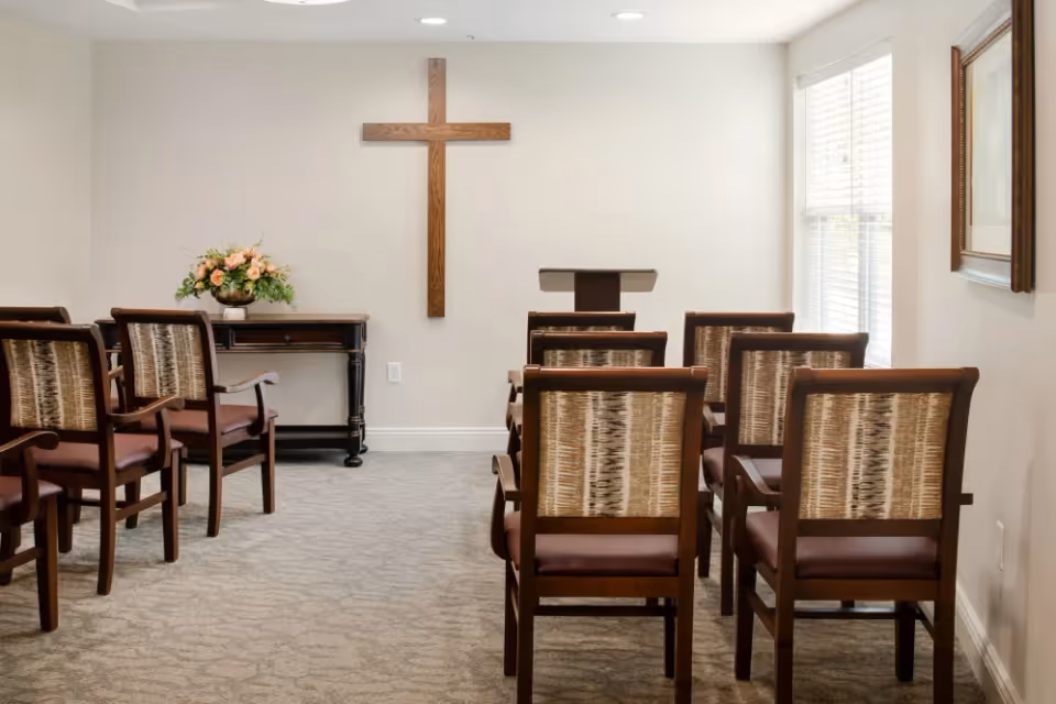 Small chapel room with wooden chairs arranged in rows facing a wooden cross mounted on a beige wall. A small table with a flower arrangement and a lectern are positioned at the front. A window with blinds and a framed picture are on the right wall.