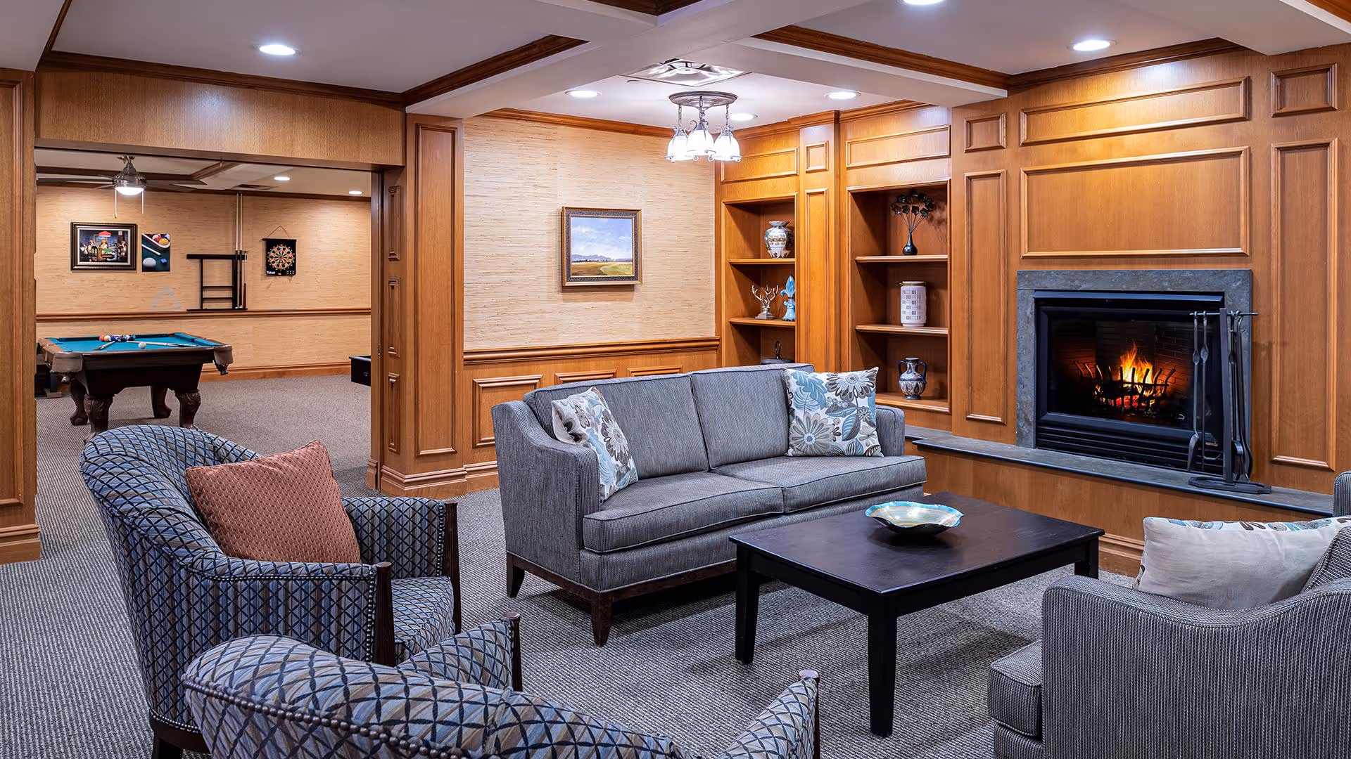 A cozy living room area with a gray sofa, two patterned armchairs, a dark wooden coffee table, and a lit fireplace set in a wooden paneled wall with built-in shelves displaying decorative items. In the background, there is a game room with a pool table and wall decorations.