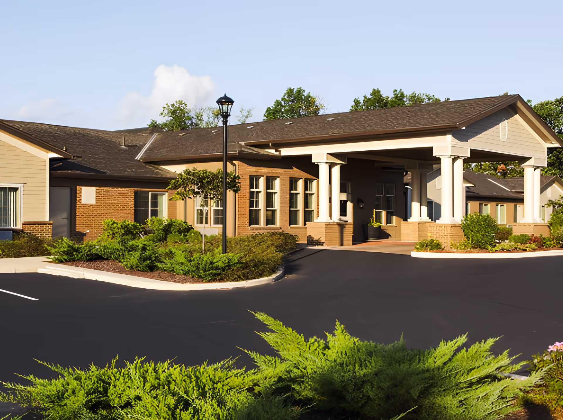 Exterior view of Juniper Glen Memory Care Assisted Living facility showing the front entrance with a covered driveway, surrounded by well-maintained bushes and greenery under a clear sky.