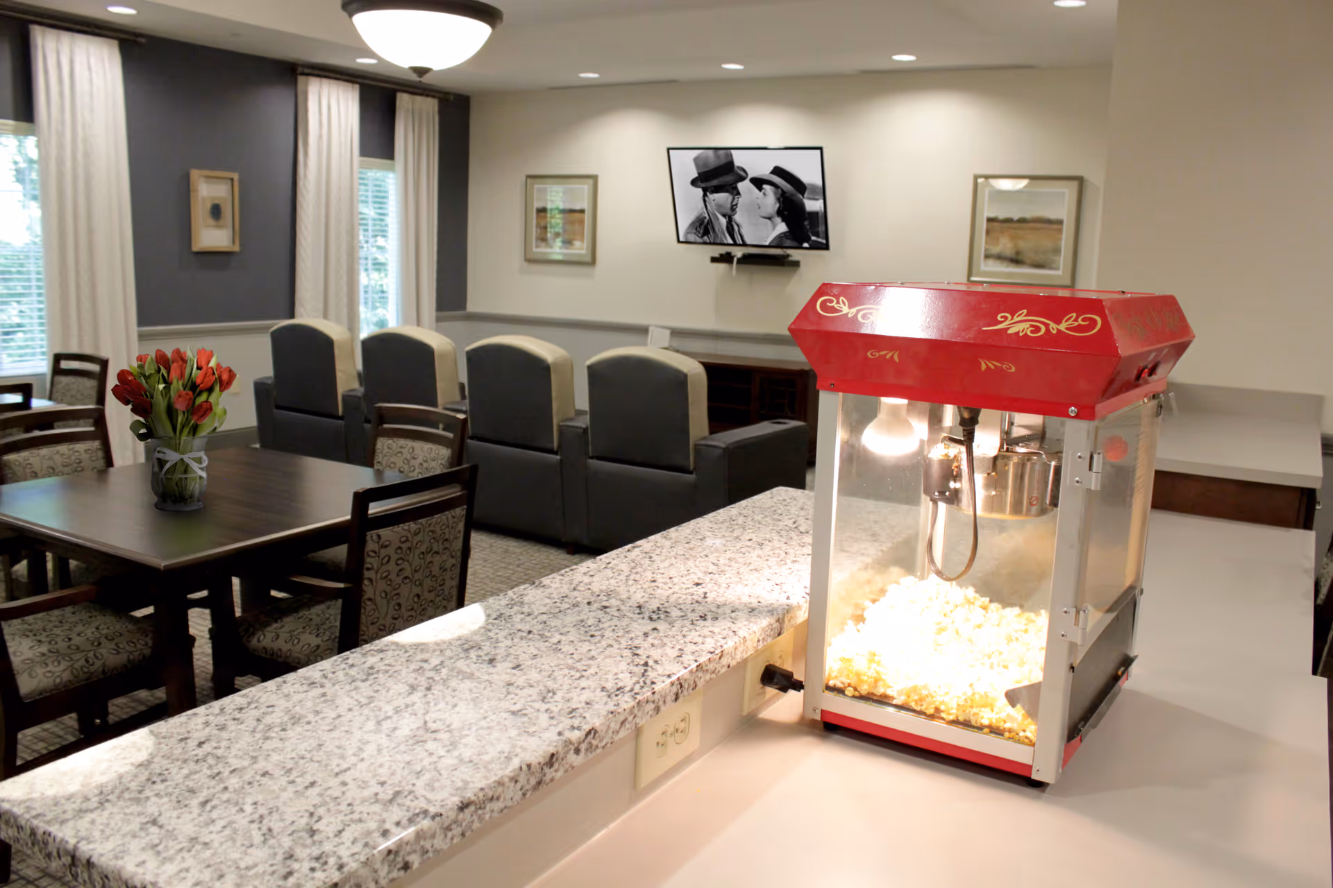 A community dining and lounge area with a popcorn machine on a countertop, a table with a vase of flowers, and armchairs facing a wall-mounted TV.