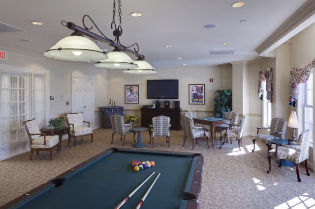 Bright common room with a pool table in the foreground, seating and game tables, and a TV on the far wall.