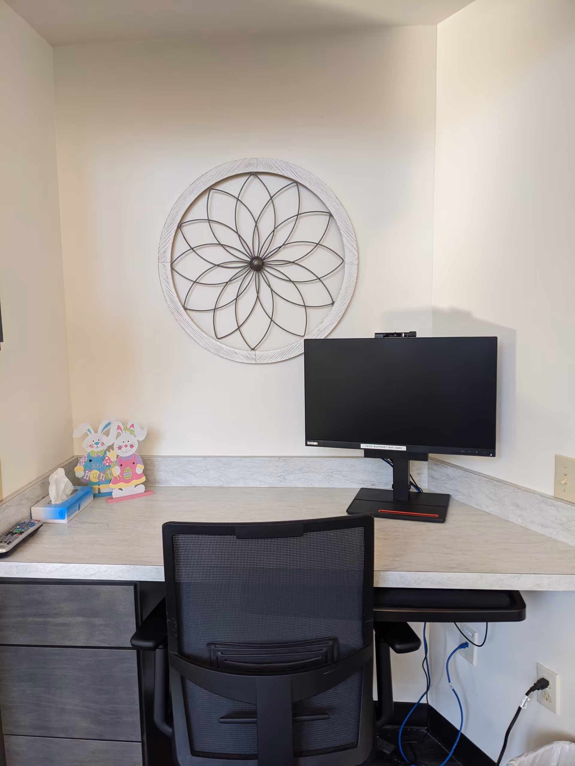 A small office or workstation with a black mesh office chair, a computer monitor on a light-colored desk, and decorative bunny figures and a tissue box on the left side of the desk. A round metal wall decoration with a floral pattern hangs on the white wall above the desk.