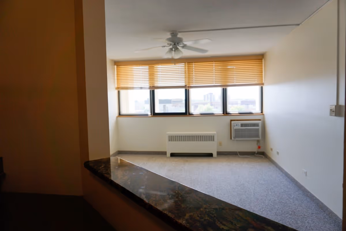 Empty room with beige walls and carpeted floor, featuring a window with wooden blinds, a ceiling fan, a radiator, and a wall-mounted air conditioning unit.