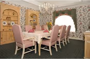 Formal dining room with a long table surrounded by upholstered pink chairs, a chandelier overhead, built-in wooden cabinets, and floral wallpaper.