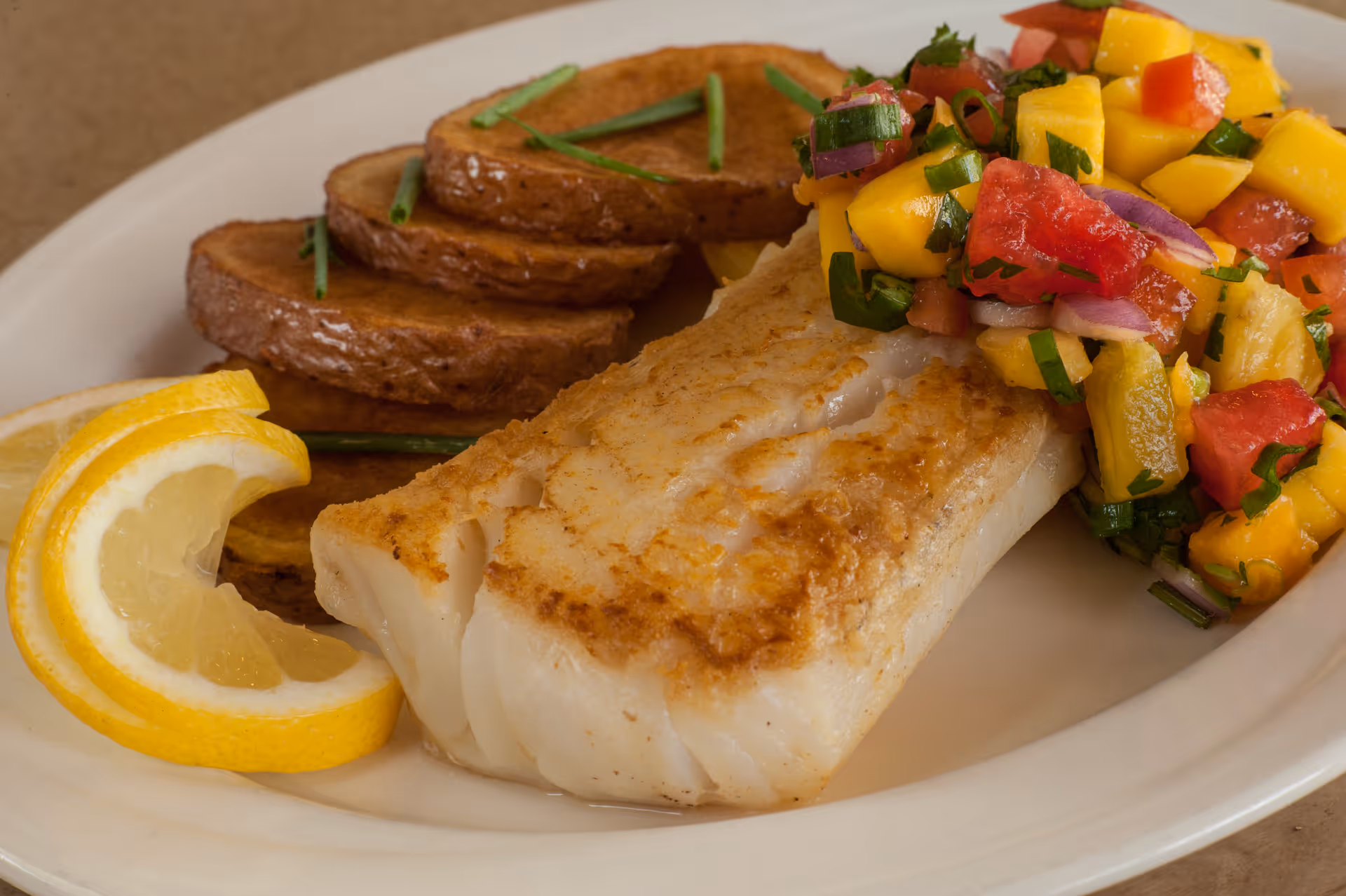 A plated meal of seared white fish with roasted potato slices, mango-tomato salsa, and lemon wedges on a white plate.