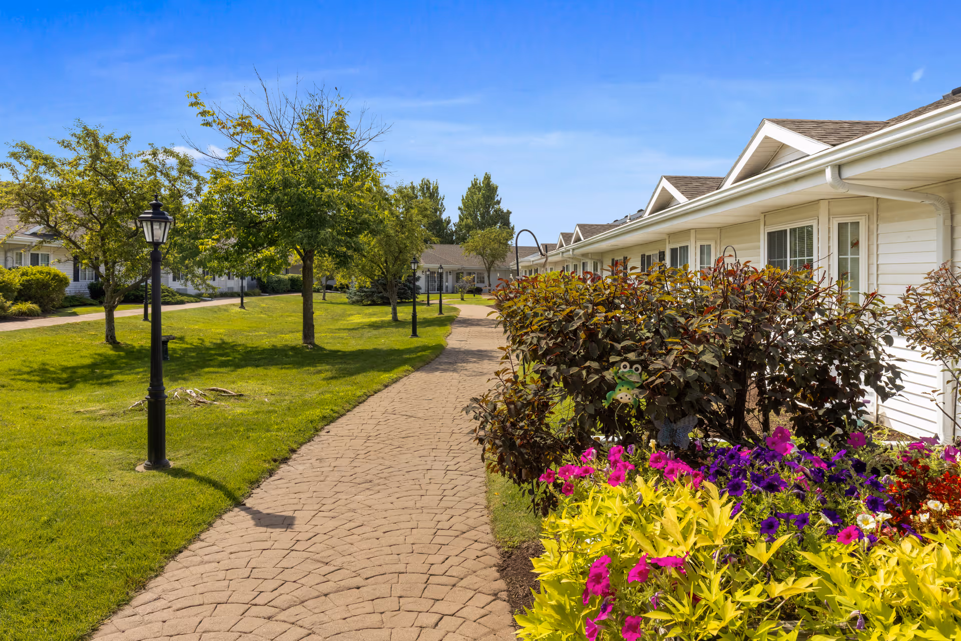 Paved walkway through a landscaped courtyard with lamp posts, flower beds, and single-story senior living buildings.
